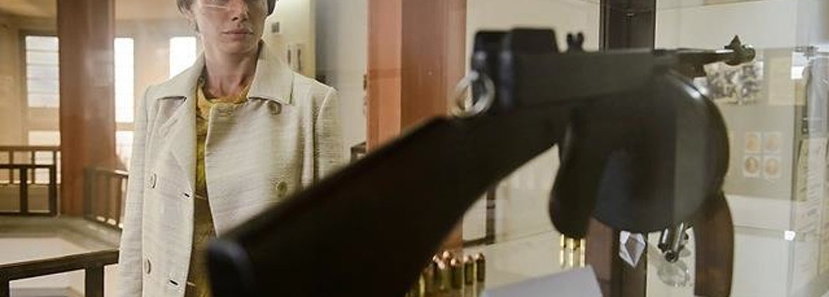 Eine in 60er Jahre Mode gekleidete Frau steht vor einer Vitrine die ein Maschinengewehr ausstellt.