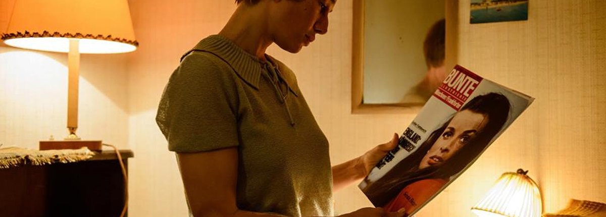 Eine junge Frau in ihrem Zimmer der 60er Jahre betrachtet ein Bunte-Magazin in atmosphärischem Licht