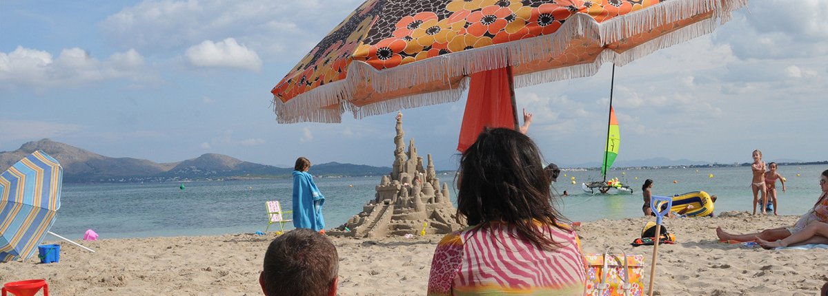 Eine Strandszene mit farbenfroh gekleideten Menschen im Vordergrund die auf eine riesige Sandburg schauen