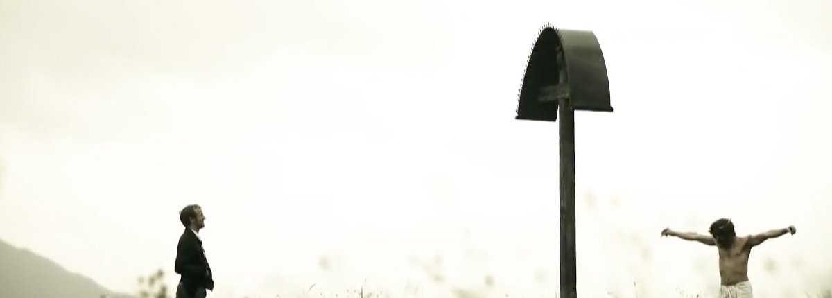 Ein Mann steht auf einer weiten Bergwiese vor einem Marterl, dahinter tanzt ein Mann mit Dornenkrone