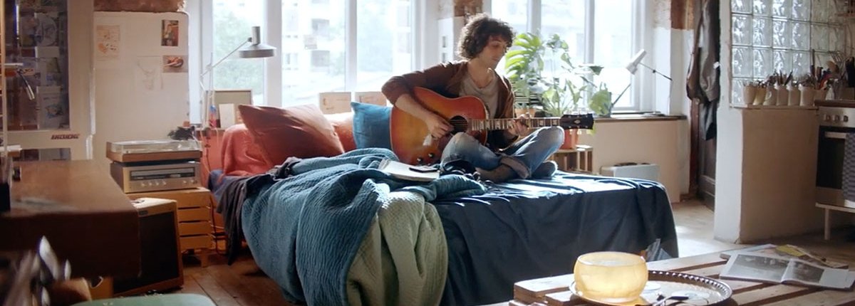 In einem lichtdurchfluteten Raum sitzt ein junger Mann auf dem Bett und spielt Gitarre
